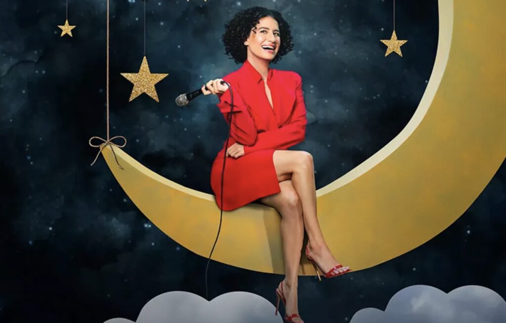 Comedian Ilana Glazer sits on a crescent moon set with a microphone in hand