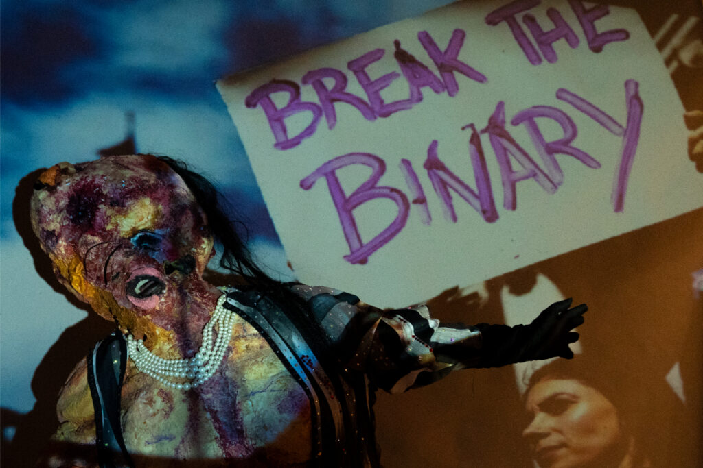 Papi Culo in their substance body suit holding a brak the binary sign. Photo by Fifth Eye Media